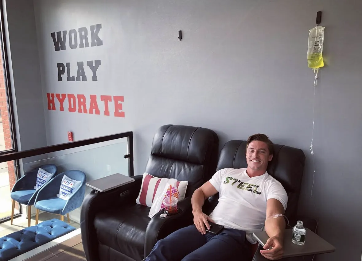 Client smiling during an IV drip beside the Work Play Hydrate wall at IV League Boston