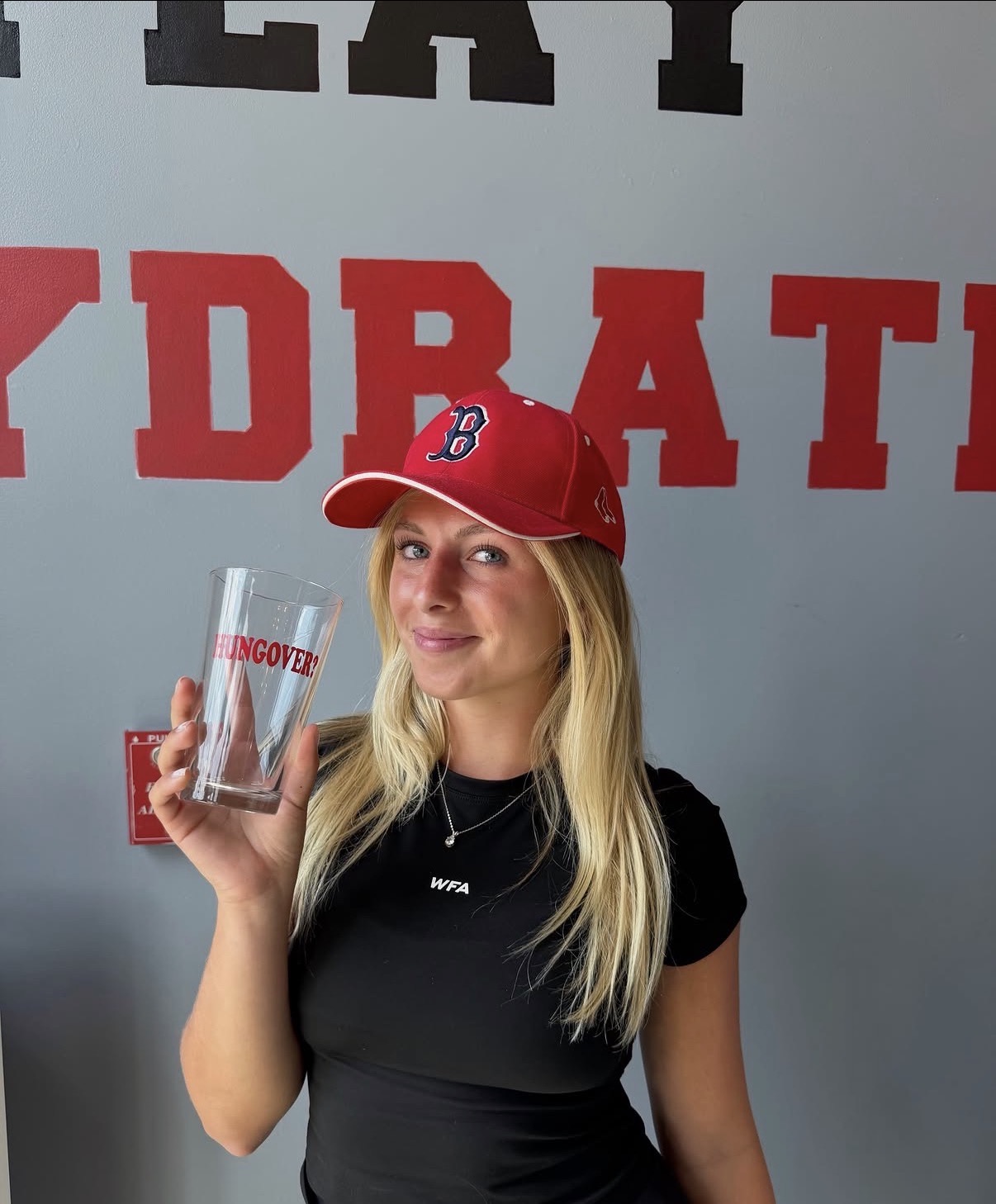 Client in a Boston Red Sox cap holding a 'Hungover?' glass before a hangover IV drip at IV League Hydration in South Boston